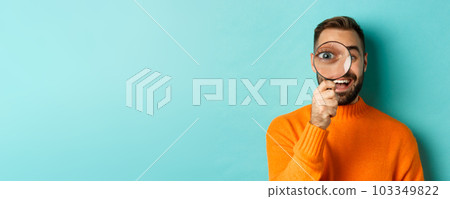 Funny man looking through magnifying glass, searching or investigating something, standing in orange sweater against turquoise background Funny man looking through magnifying glass, searching or investigating something, standing in orange sweater against turquoise background 103349822