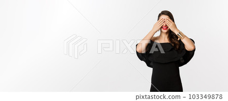 Fashion and beauty. Beautiful woman in black dress and red lipstick, covering eyes and smiling, waiting for surprise, standing over white background Fashion and beauty. Beautiful woman in black dress and red lipstick, covering eyes and smiling, waiting for surprise, standing over white background 103349878