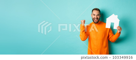 Real estate concept. Excited man saying yes, showing paper house maket and looking satisfied, standing in orange sweater over light blue background 103349916