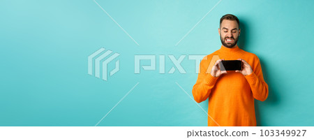 Image of man cringe at something on mobile screen, staring disgusted at display, showing smartphone, standing over light blue background Image of man cringe at something on mobile screen, staring disgusted at display, showing smartphone, standing over light blue background 103349927