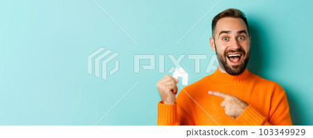 Real estate. Excited bearded man pointing at small paper house maket, standing amused over light blue background 103349929