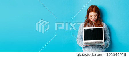 Cute redhead woman in sweater, showing and looking at laptop screen with pleased smile, demonstrating something online, standing over blue background 103349936