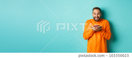 Handsome adult man celebrating birthday, blowing out candle on cake and making wish, standing against turquoise background 103350023