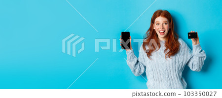 Excited redhead girl showing mobile phone screen and credit card, demonstrating online store or application, standing over blue background Excited redhead girl showing mobile phone screen and credit card, demonstrating online store or application, standing over blue background 103350027