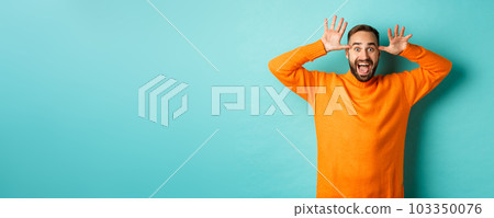 Image of handsome caucasian man making funny faces, mocking someone and smiling, standing against light blue background Image of handsome caucasian man making funny faces, mocking someone and smiling, standing against light blue background 103350076