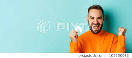 Real estate. Cheerful man buying apartment, rejoicing and saying yes, showing small paper house, standing over light blue background 103350083