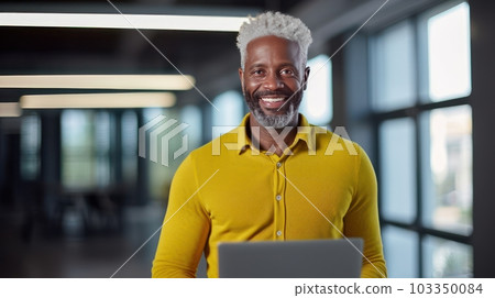 Portrait of a dark-skinned adult man with gray hair, who smiles and sits in the office at a laptop in a bright office, wearing a yellow shirt. AI generated. 103350084