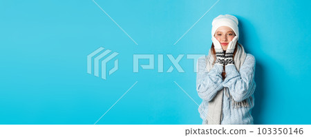 Winter and holidays concept. Cute girl in white beanie, sweater and gloves, squeezing cheeks and smiling pleased, warm-up after cold outdoors, standing over blue background Winter and holidays concept. Cute girl in white beanie, sweater and gloves, squeezing cheeks and smiling pleased, warm-up after cold outdoors, standing over blue background 103350146