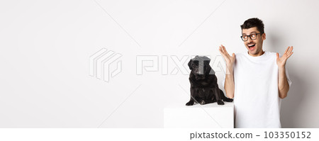 Image of cheerful young man rejoicing, looking at cute black dog pug and smiling, standing over white background 103350152