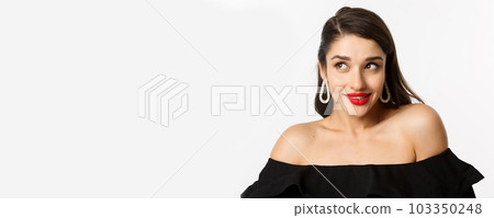 Fashion and beauty concept. Close-up of beautiful woman with red lips, makeup and black dress, showing her earrings and looking pleased, white background Fashion and beauty concept. Close-up of beautiful woman with red lips, makeup and black dress, showing her earrings and looking pleased, white background 103350248