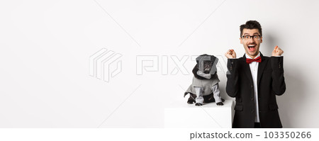 Animals, party and celebration concept. Happy young man in suit and puppy in pet cosume standing over white background, dog owner rejoicing and triumphing Animals, party and celebration concept. Happy young man in suit and puppy in pet cosume standing over white background, dog owner rejoicing and triumphing 103350266