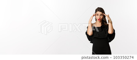 Fashion and beauty. Annoyed and tired woman in black dress, touching head and roll eyes bothered, standing distressed against white background Fashion and beauty. Annoyed and tired woman in black dress, touching head and roll eyes bothered, standing distressed against white background 103350274