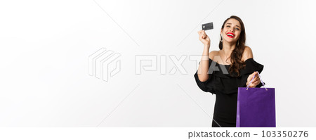 Stylish young woman in black dress going shopping, holding bag and credit card, smiling pleased, standing over white background Stylish young woman in black dress going shopping, holding bag and credit card, smiling pleased, standing over white background 103350276