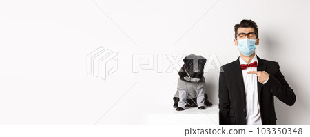 Coronavirus, pets and celebration concept. Disappointed young man in face mask and suit, pointing finger at cute black pug dog wearing party costume, standing over white background 103350348