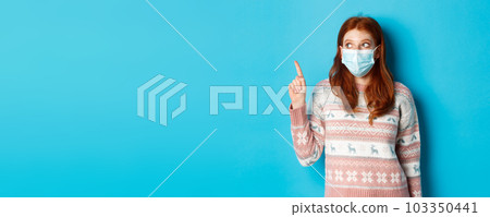 Winter, covid-19 and quarantine concept. Curious redhead girl in medical mask picking product, looking and pointing at upper left corner promo, blue background 103350441