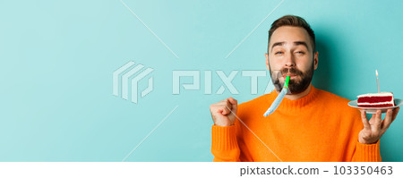 Close-up of funny adult man celebrating his birthday, holding bday cake with candle, blowing party wistle and rejoicing, standing over light blue background 103350463