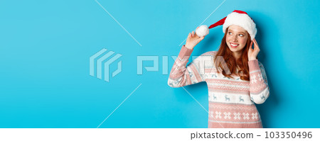 Winter and Christmas Eve concept. Cute teenage girl wearing santa hat and sweater, celebrating xmas, smiling happy, standing over blue background 103350496