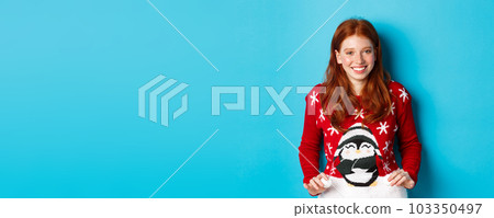 Winter holidays and Christmas Eve concept. Cute redhead girl wearing adorable sweater and smiling, celebrating New Year, standing over blue background 103350497