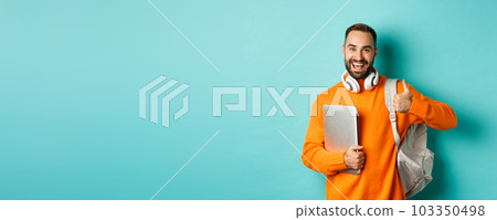Happy man with backpack and headphones, holding laptop and smiling, showing thumb-up in approval, standing over turquoise background 103350498