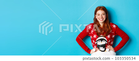 Winter holidays and Christmas Eve concept. Beautiful teenage redhead girl in xmas sweater looking at camera, smiling carefree, holding hands on waist, blue background 103350534
