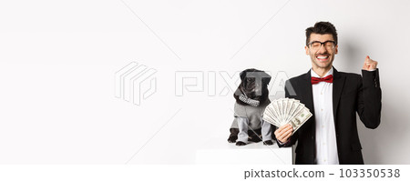 Happy young man and cute black dog standing in party costumes, pug owner holding money dollars and rejoicing, winning prize, white background Happy young man and cute black dog standing in party costumes, pug owner holding money dollars and rejoicing, winning prize, white background 103350538