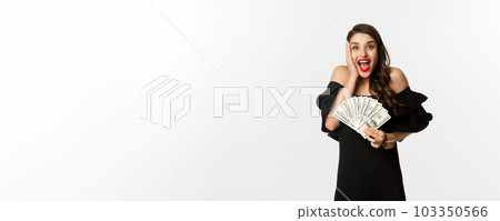 Fashion and shopping concept. Attractive woman rejoicing of money prize, holding dollars and shouting from excitement, standing in black dress over white background 103350566