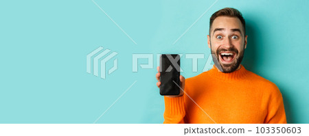 Close-up of handsome bearded guy in orange sweater, showing smartphone screen and smiling, showing promo online, turquoise background 103350603