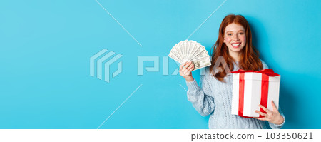 Young redhead woman holding Christmas gift box and money, smiling pleased, standing over blue background 103350621