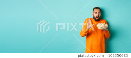 Portrait of scared guy holding bowl of popcorn and declining something, showing stop gesture, standing over blue background Portrait of scared guy holding bowl of popcorn and declining something, showing stop gesture, standing over blue background 103350683