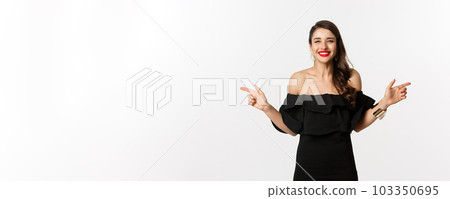 Fashion and beauty. Attractive woman in jewelry, makeup and black dress, smiling and pointing fingers sideways copy space offer, white background 103350695