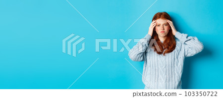 Distressed teenage redhead girl touching head, looking with troubled face expression, standing against blue background, have problem 103350722