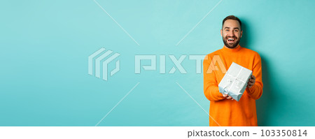 Holidays and celebration concept. Cheerful bearded man giving present, holding gift and smiling, congratulating you, standing over turquoise background Holidays and celebration concept. Cheerful bearded man giving present, holding gift and smiling, congratulating you, standing over turquoise background 103350814