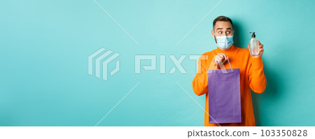 Coronavirus, pandemic and lifestyle concept. Man in face mask showing shopping bag and hand sanitizer, standing over turquoise background 103350828