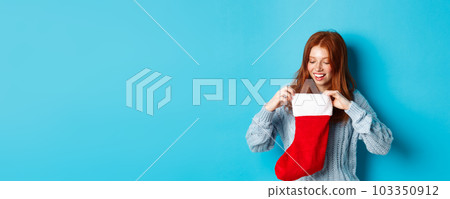 Winter holidays and gifts concept. Funny redhead girl looking inside Christmas stocking and smiling happy, receiving xmas present, standing against blue background Winter holidays and gifts concept. Funny redhead girl looking inside Christmas stocking and smiling happy, receiving xmas present, standing against blue background 103350912