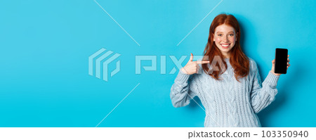 Satisfied redhead girl pointing at phone screen, showing smartphone app or online promo and smiling, standing in sweater against blue background Satisfied redhead girl pointing at phone screen, showing smartphone app or online promo and smiling, standing in sweater against blue background 103350940