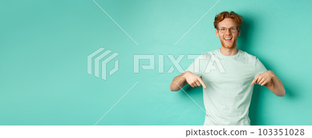 Happy young man with red hair, wearing glasses and t-shirt, showing advertisement, smiling and pointing fingers down, standing over turquoise background 103351028