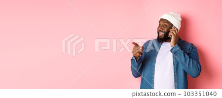 Handsome modern Black man talking on mobile phone, pointing left at person and smiling, standing over pink background 103351040