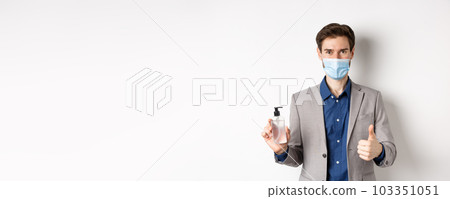 Covid-19, pandemic and business concept. Businessman in office suit and medical mask showing bottle of hand sanitizer and thumb up, recommend use antiseptic at work 103351051
