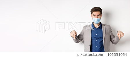 Covid-19, pandemic and business concept. Hesitant man in suit and medical mask asking opinion, pointing fingers down and look at camera, white background 103351064