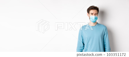 Covid-19, pandemic and social distancing concept. Cheerful caucasian man in face mask winking at camera, looking happy, standing over white background 103351172