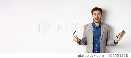 Online shopping. Surprised businessman holding plastic credit card and smartphone, making money in internet, standing in suit against white background 103351228