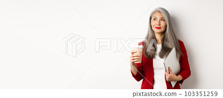 Business. Successful asian businesswoman with grey hair, drinking coffee and standing with laptop, looking thoughtful upper left corner 103351259