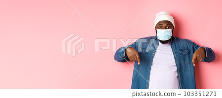 Covid-19, lifestyle and lockdown concept. Handsome african-american man in face mask, pointing fingers down, showing promo offer on bottom, standing over pink background Covid-19, lifestyle and lockdown concept. Handsome african-american man in face mask, pointing fingers down, showing promo offer on bottom, standing over pink background 103351271