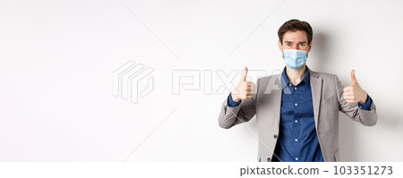 Covid-19, pandemic and business concept. Cheerful man in suit and medical mask using preventive measures in office, showing thumbs up, white background 103351273