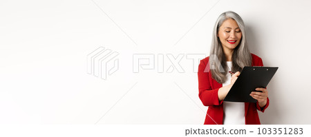 Successful asian senior businesswoman with grey hair taking notes on clipboard, inspecting enterprise, standing over white background Successful asian senior businesswoman with grey hair taking notes on clipboard, inspecting enterprise, standing over white background 103351283