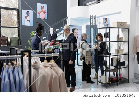 African american employee helping client with formal outfit, checking tie fabric in clothing store. Senior customer man buying trendy casual wear and fashionable accessories in modern boutique African american employee helping client with formal outfit, checking tie fabric in clothing store. Senior customer man buying trendy casual wear and fashionable accessories in modern boutique 103351643