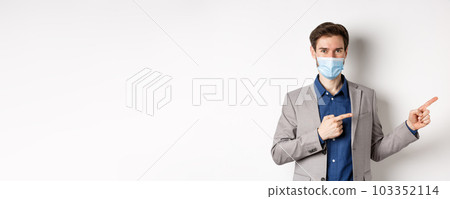 Covid-19, pandemic and business concept. Confident male manager in suit and medical mask showing way, pointing fingers right at advertisement, white background Covid-19, pandemic and business concept. Confident male manager in suit and medical mask showing way, pointing fingers right at advertisement, white background 103352114