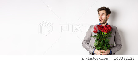 Handsome young man in suit holding red roses, looking left with surprised and startled expression, standing on Valentines day over white background Handsome young man in suit holding red roses, looking left with surprised and startled expression, standing on Valentines day over white background 103352125