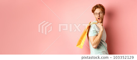 Carefree young man with red hair and glasses, walking with shopping bag, looking behind his shoulder with amazed expression, standing over pink background 103352129