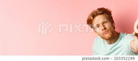 Funny young man with red messy hair taking selife with puckered lips and macho expression, view from smartphone camera, standing over pink background 103352299